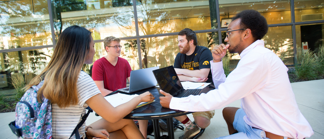 In this photo a group of four Iowa students, 3 boys one girl, sit around an outdoor table with laptops as they study in front of Blank Honors Center