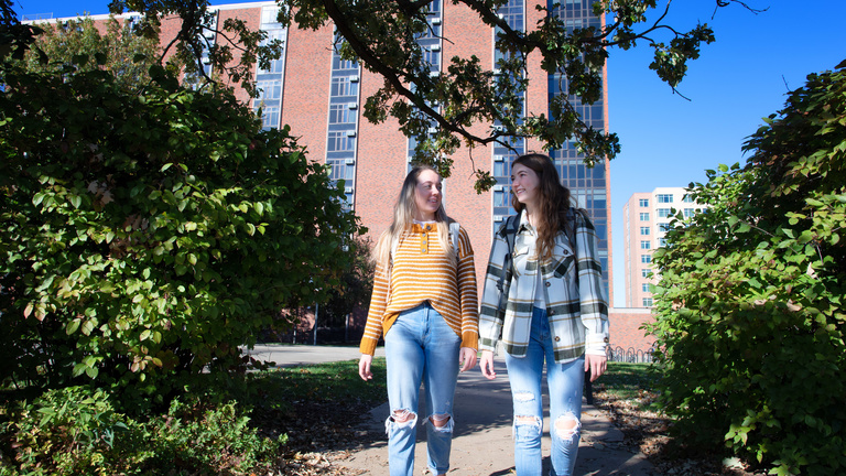 Two female students walking on a sidewalk and talking with one another