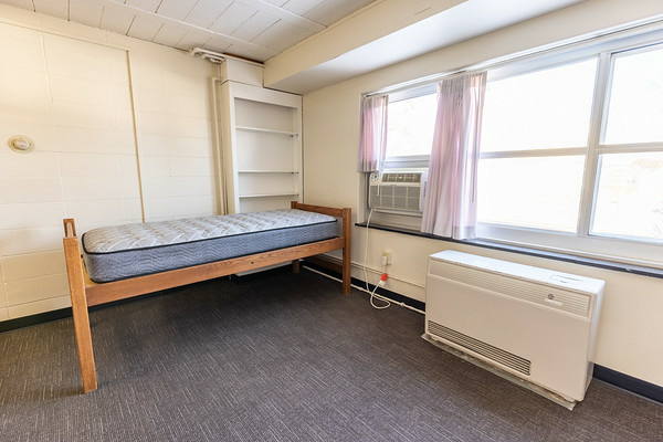 a Parklawn studio, shown with bed against big set of windows, air conditioning and radiator, with a built-in bookshelf in the corner.