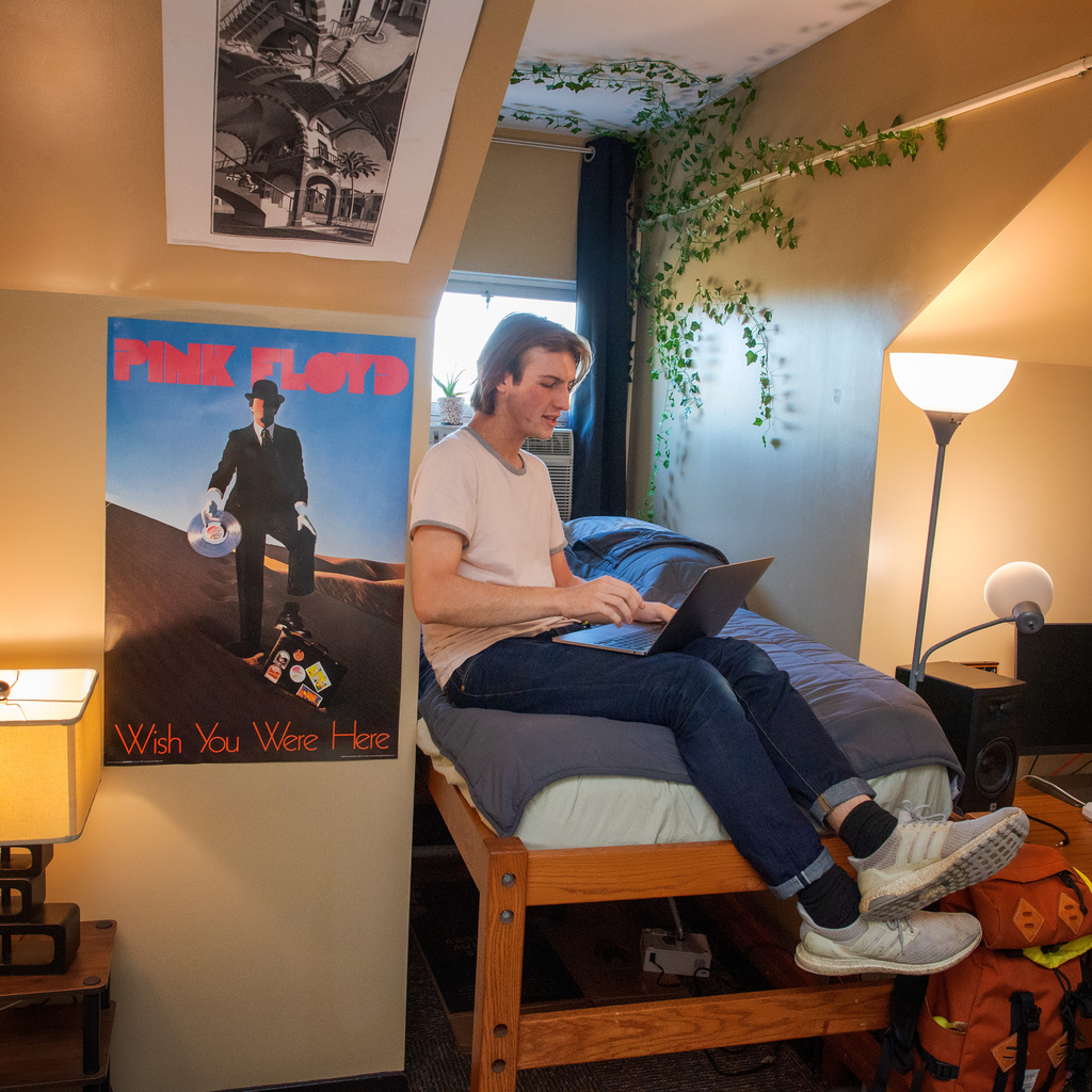 Student sitting on their bed and typing on a laptop with Pink Floyd band poster in background