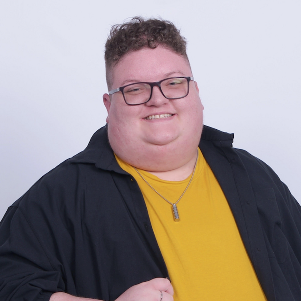 pictured is a person with curly brown hair on top, cropped short on the sides, wearing glasses and a yellow t shirt under a dark gray jacket