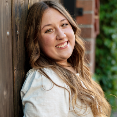 headshot of Aubrey Stark, a woman wearing a white shirt with puffed sleeves, leaning against a wooden door