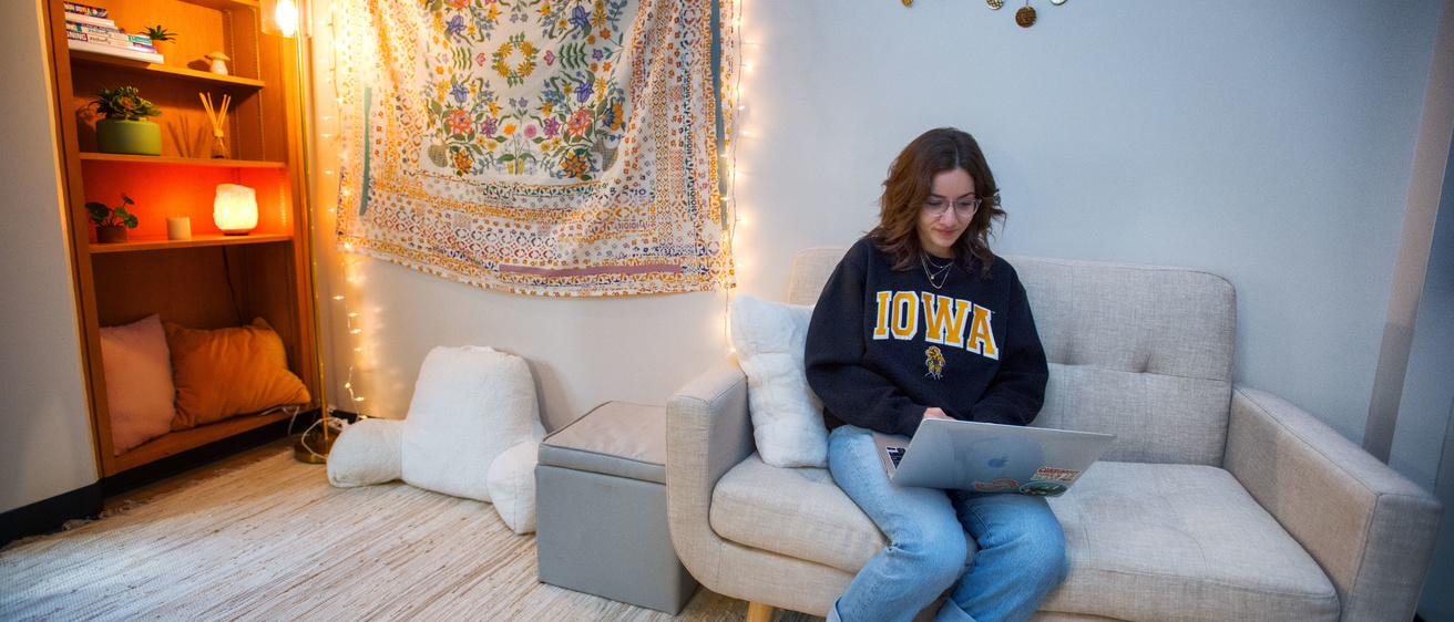 Student sitting on a white couch typing on a laptop and wearing a sweatshirt