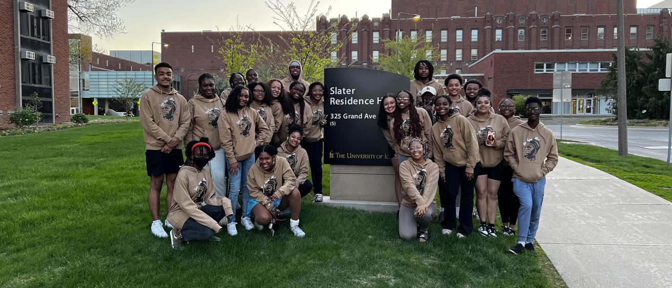 Members of the Young, Gifted, and Black gather on the lawn in front of their residence hall wearing matching brown sweatshirts with an Iowa and YGB design