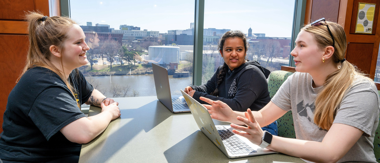 three students sit at a booth in Hillcrest overlooking the river, talking and working on their laptops