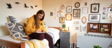 Student sitting on a bed writing <a href=